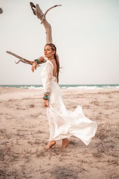 Model in boho style in a white long dress and silver jewelry on the beach. Her hair is braided, and there are many bracelets on her arms