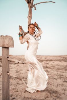 Model in boho style in a white long dress and silver jewelry on the beach. Her hair is braided, and there are many bracelets on her arms