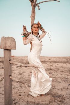 Model in boho style in a white long dress and silver jewelry on the beach. Her hair is braided, and there are many bracelets on her arms