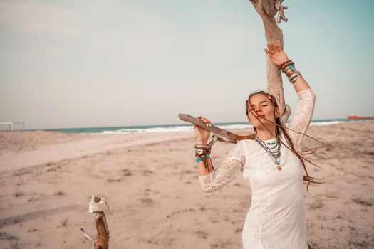 Model in boho style in a white long dress and silver jewelry on the beach. Her hair is braided, and there are many bracelets on her arms