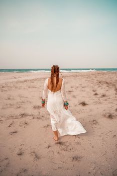 Model in boho style in a white long dress and silver jewelry on the beach. Her hair is braided, and there are many bracelets on her arms