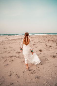 Model in boho style in a white long dress and silver jewelry on the beach. Her hair is braided, and there are many bracelets on her arms