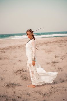 Model in boho style in a white long dress and silver jewelry on the beach. Her hair is braided, and there are many bracelets on her arms