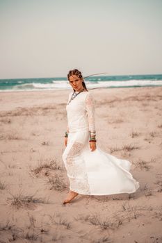 Model in boho style in a white long dress and silver jewelry on the beach. Her hair is braided, and there are many bracelets on her arms