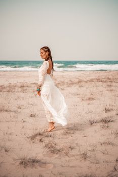 Model in boho style in a white long dress and silver jewelry on the beach. Her hair is braided, and there are many bracelets on her arms