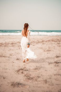 Model in boho style in a white long dress and silver jewelry on the beach. Her hair is braided, and there are many bracelets on her arms