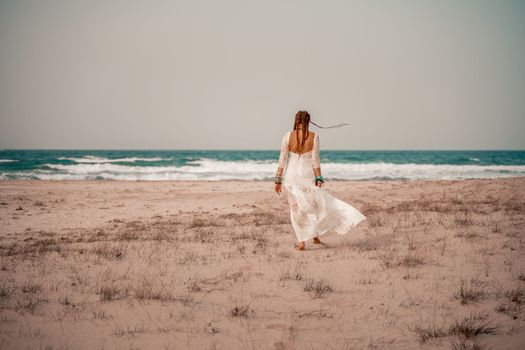 Model in boho style in a white long dress and silver jewelry on the beach. Her hair is braided, and there are many bracelets on her arms