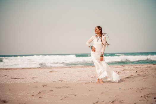 Model in boho style in a white long dress and silver jewelry on the beach. Her hair is braided, and there are many bracelets on her arms