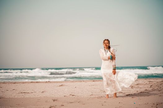 Model in boho style in a white long dress and silver jewelry on the beach. Her hair is braided, and there are many bracelets on her arms