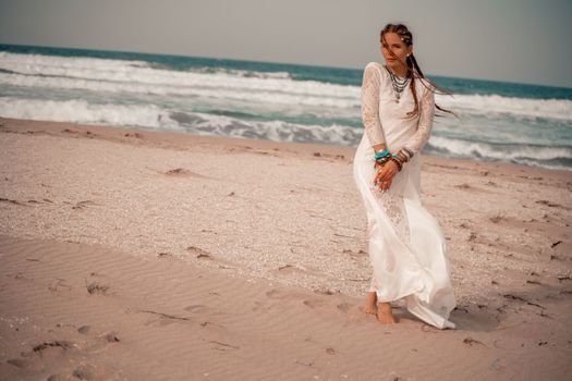 Model in boho style in a white long dress and silver jewelry on the beach. Her hair is braided, and there are many bracelets on her arms