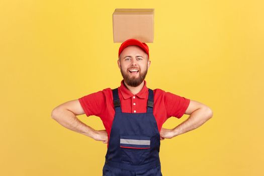 Portrait of funny courier man wearing blue uniform standing with hands on hips and holding cardboard box on his head, looking smiling at camera. Indoor studio shot isolated on yellow background.