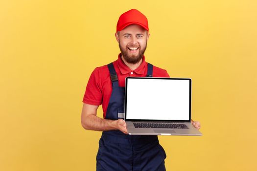 Portrait of smiling happy worker man in uniform standing and showing laptop with blank screen for advertisement, looking smiling at camera. Indoor studio shot isolated on yellow background.