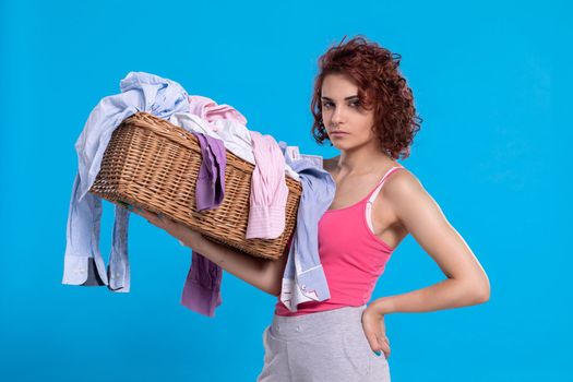 A woman with an angry face carries a wicker basket with shirts for washing. Daily household chores. Young attractive girl with curly hair.