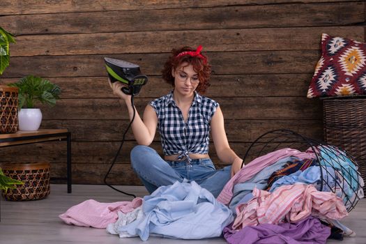 A young girl is waiting to iron men's shirts. She sits on the floor of a room in the house. An overturned basket of clothes. A wall of brown boards.
