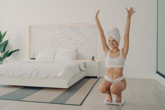 Young satisfied beautiful lady in perfect shape squatted on scales in white underwear and towel wrapped on head after morning shower in bedroom, throwing her hands up celebrating her weight loss