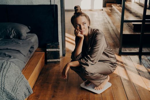 Upset and unmotivated woman on electronic floor scale in bedroom wondering why cannot lose weight despite her new diet, looking aside with sade face expression. Weight Loss and healthy lifestyle