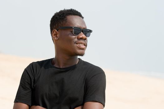 portrait of young man in black t-shirt standing arms crossed with black glasses and smiling at the beach.