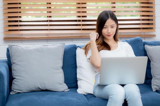 Young asian business woman smile and work from home with laptop computer online to internet on sofa in living room, freelance girl using notebook on couch with comfort, new normal, lifestyle concept.