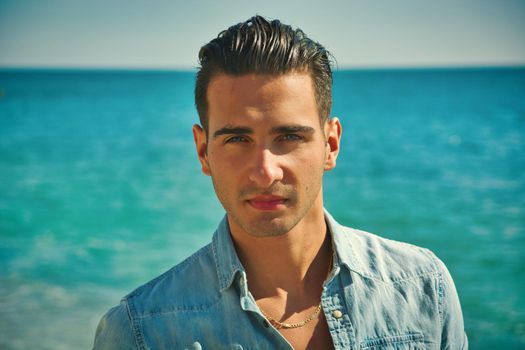 Three Quarter Shot of a Handsome Athletic Young Man in Trendy Attire, on a Beach in a Sunny Summer Day, Looking At Camera against Blue Sky Background.
