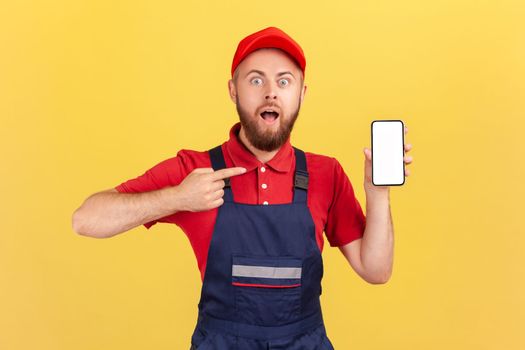 Portrait of surprised repairman in blue overalls red t-shirt pointing finger at smartphone with empty display, online service order app. Indoor studio shot isolated on yellow background.