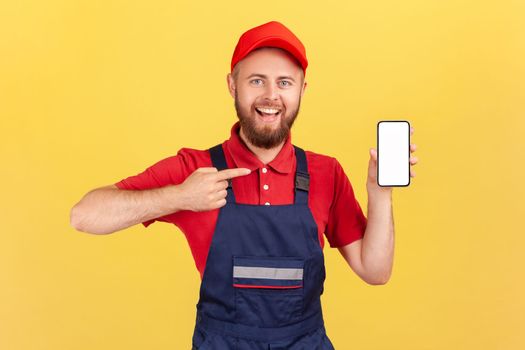 Portrait of joyful repairman in blue overalls red t-shirt pointing finger at smartphone with empty display, online service order app. Indoor studio shot isolated on yellow background.
