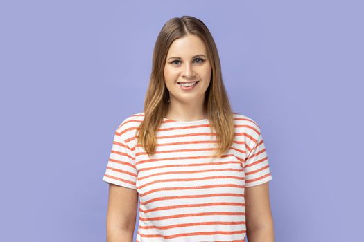 Portrait of delighted smiling blond woman wearing striped T-shirt standing looking at camera with happy facial expression, being in good mood. Indoor studio shot isolated on purple background.