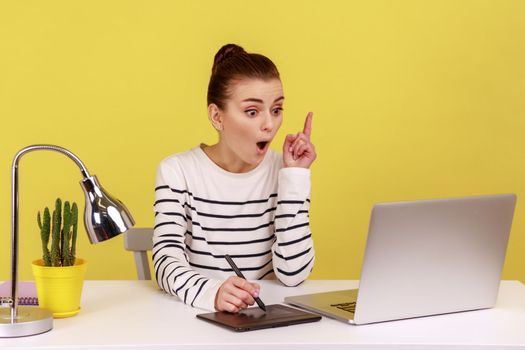 Excited woman designer pointing finger up, having idea, drawing with stylus on graphic tablet, sitting in office working on new collection. Indoor studio studio shot isolated on yellow background.