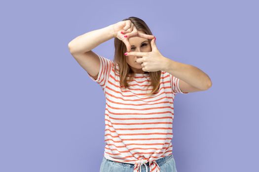 Blond woman wearing striped T-shirt gesturing picture frame with hands, looking through fingers and focusing on interesting moment, taking photo. Indoor studio shot isolated on purple background.