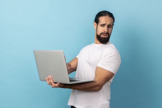 Portrait of handsome man with beard wearing white T-shirt turning his face from display, cause dont want to see in the laptop screen. Indoor studio shot isolated on blue background.