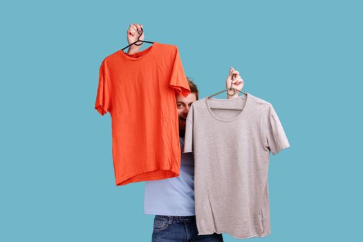 Portrait of optimistic young adult handsome bearded man peeping from T-shorts on hangers, choosing attire, expressing positive emotions. Indoor studio shot isolated on blue background.