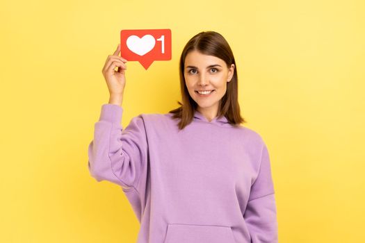 Social media Like button. Portrait of happy woman raised arm and holding heart icon, recommending to follow and share blog with trendy content. Indoor studio shot isolated on yellow background.