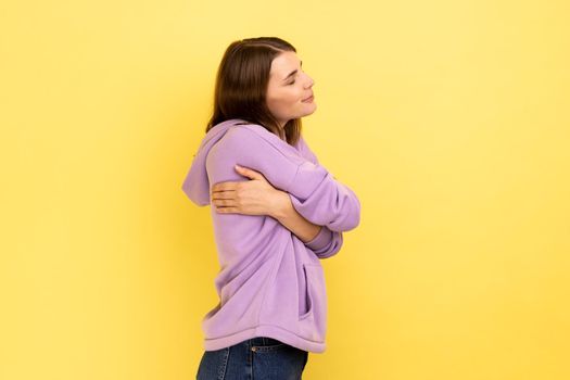 I love myself. Side view of selfish narcissistic young woman embracing herself and smiling with dreamy satisfied expression, wearing purple hoodie. Indoor studio shot isolated on yellow background.