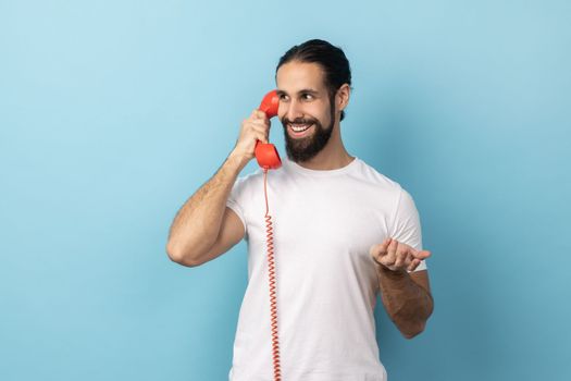 Portrait of positive man with beard wearing white T-shirt talking on red retro telephone, having pleasant conversation, looking away with smile Indoor studio shot isolated on blue background.