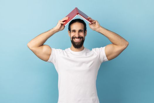 Portrait of delighted man with beard wearing white T-shirt making roof, holding book over head, looking at camera with happiness. Indoor studio shot isolated on blue background.