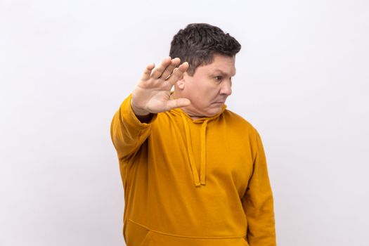 Portrait of middle aged man showing stop sign, standing with turning face, meaning caution to avoid danger or mistake, wearing urban style hoodie. Indoor studio shot isolated on white background.