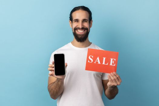Portrait of handsome smiling man with beard wearing white T-shirt holding sale card and smartphone, being satisfied with online sell-out. Indoor studio shot isolated on blue background.