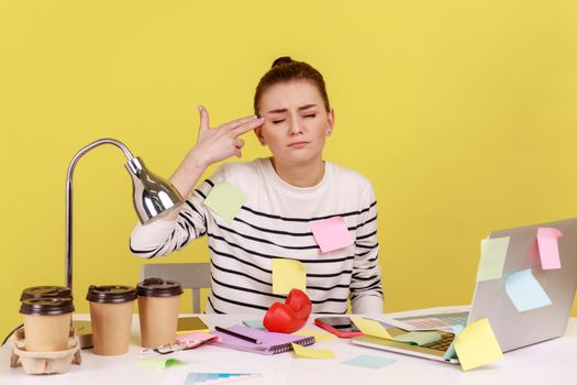 Depressed woman covered with sticky notes working on laptop, pointing finger gun gesture to head, committing suicide from stressful job. Indoor studio studio shot isolated on yellow background.