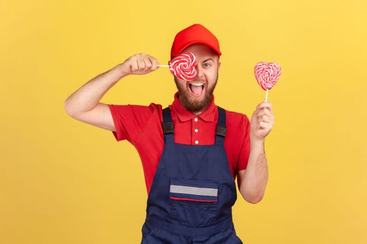 Childish playful worker man wearing red cap and blue uniform holding tasty sugary candy and covering eye with heart shape lollypop. Indoor studio shot isolated on yellow background.