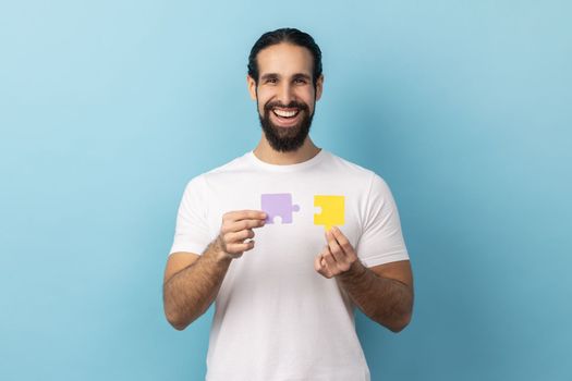 Portrait of man with beard wearing white T-shirt holding yellow and purple puzzle pieces, solving tasks, looking at camera with toothy smile. Indoor studio shot isolated on blue background.