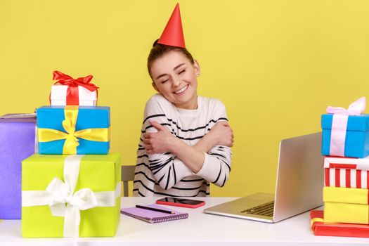 Satisfied woman office employee with party cone sitting at workplace surrounded by many presents and embracing herself, celebrating birthday. Indoor studio studio shot isolated on yellow background.