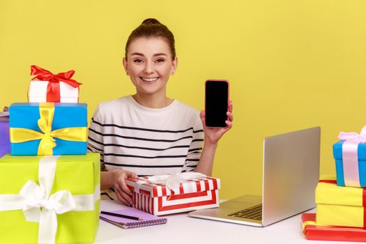 Happy woman office manager sitting at workplace, holding gift box and cell phone with blank display, empty mock up screen on mobile device. Indoor studio studio shot isolated on yellow background.