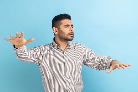 Loneless and blindness. Lost worry bearded businessman standing with closed eyes and try to touching something or find, wearing striped shirt. Indoor studio shot isolated on blue background.