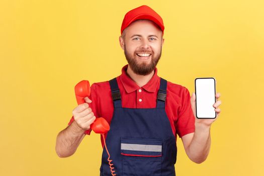 Portrait of delighted smiling worker man holding out retro phone and cell phone with empty screen for advertisement, choose best device for you, Indoor studio shot isolated on yellow background.