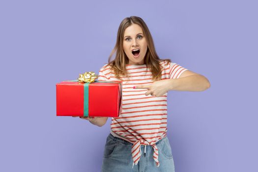 Portrait of astonished surprised blond woman wearing striped T-shirt holding and pointing red present box, presenting gift for holiday. Indoor studio shot isolated on purple background.