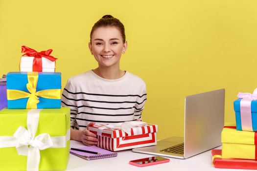 Happy woman office manager sitting at workplace surrounded by many presents, smiling and holding gift box, celebrating professional holiday. Indoor studio studio shot isolated on yellow background.