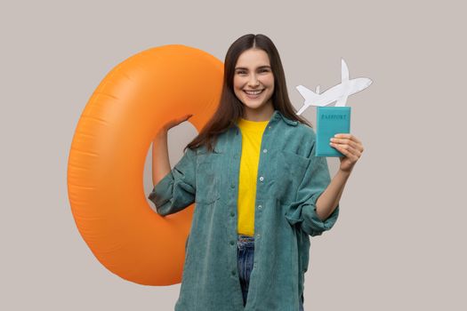 Portrait of joyful dark haired woman holding orange rubber ring, passport document and paper airplane, traveling abroad, wearing casual style jacket. Indoor studio shot isolated on gray background.