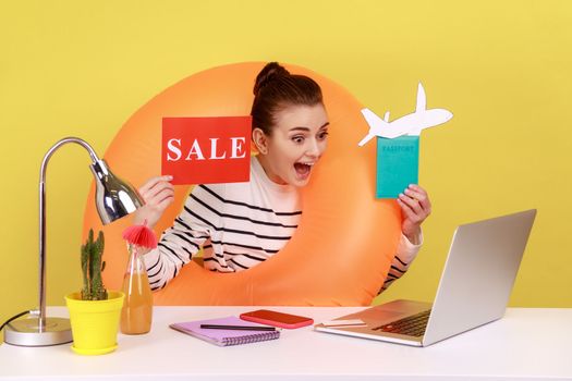 Excited amazed woman with rubber ring, holding card with sale inscription, passport and paper plane, having video call. Indoor studio studio shot isolated on yellow background.