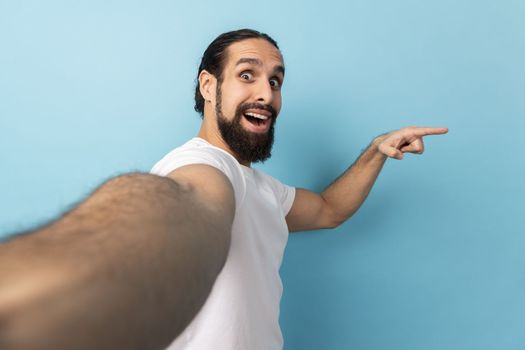 Portrait of excited man wearing white T-shirt looking at camera with positive expression, pointing aside at copy space, POV, point of view of photo. Indoor studio shot isolated on blue background.