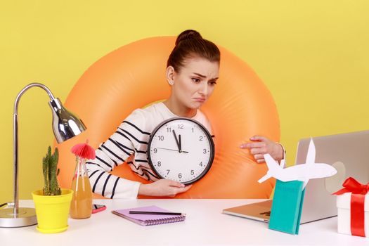 Sad upset woman in rubber ring on neck, showing wall clock to laptop screen, has video call, has no time to go on summer vacation. Indoor studio studio shot isolated on yellow background.
