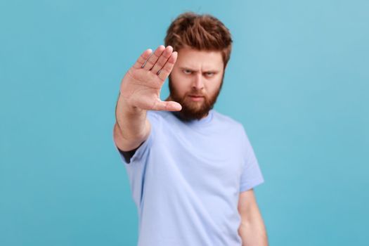 Portrait of strict bearded man showing stop sign at camera, standing with frowning face and bossy expression, meaning caution to avoid danger or mistake. Indoor studio shot isolated on blue background